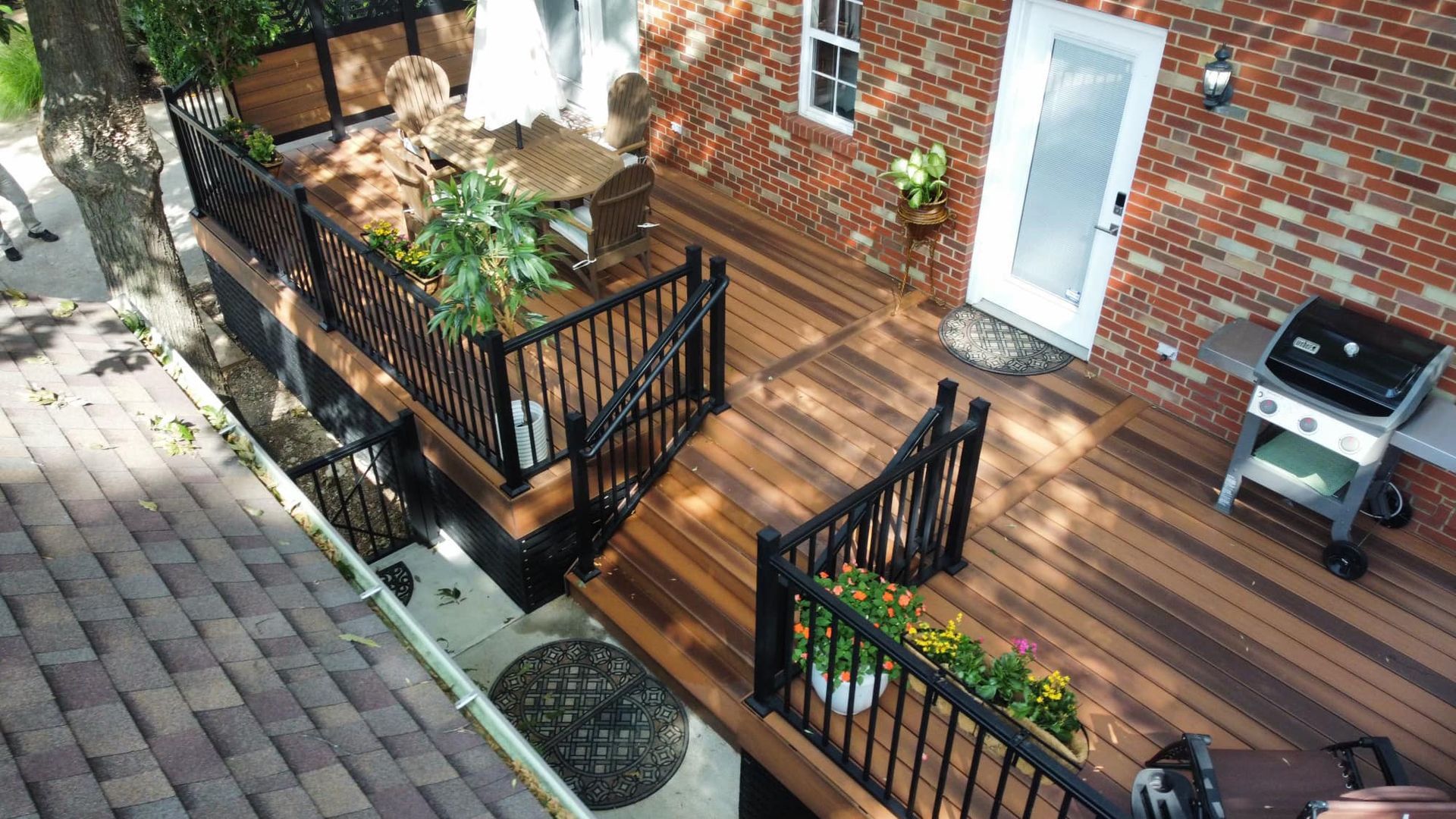 Elevated wooden deck with black railings and stairs. Adjacent to a brick building with a grill.