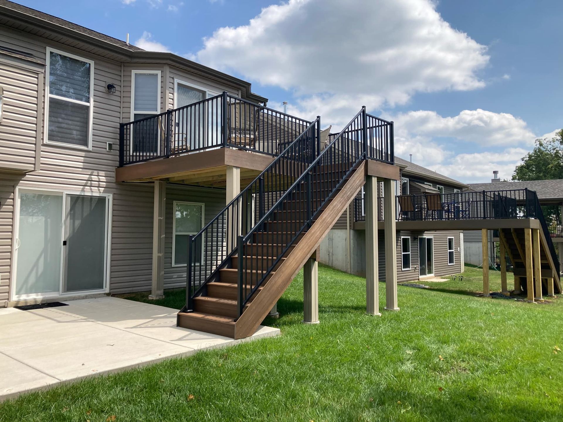 Backyard with a two-level deck, dark railing, and stairs. Green grass, blue sky.