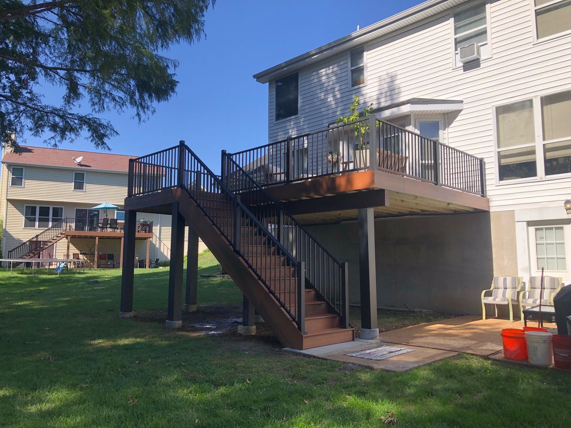 Backyard deck with stairs. Brown deck and stairs, black railing, attached to a two-story beige house. Sunny day.