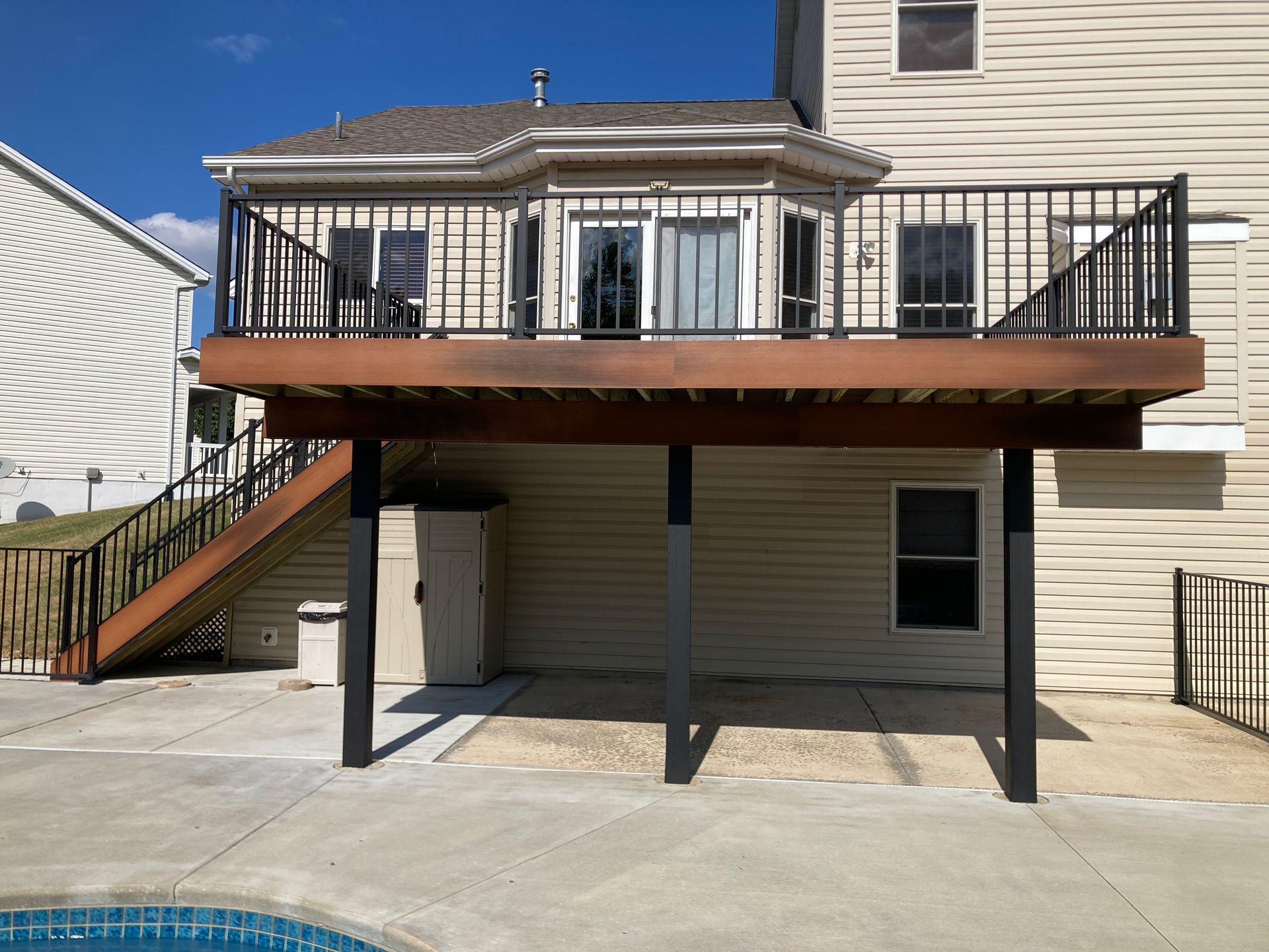 Deck with black railing, overlooking a pool. Brown deck, tan siding, blue sky. Stairs on the left.