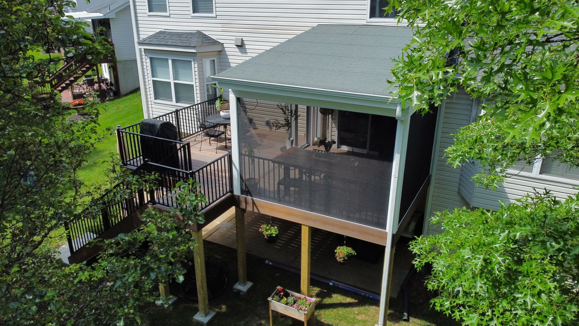 Screened-in deck attached to a house with a grill and hanging flower baskets; surrounded by trees.
