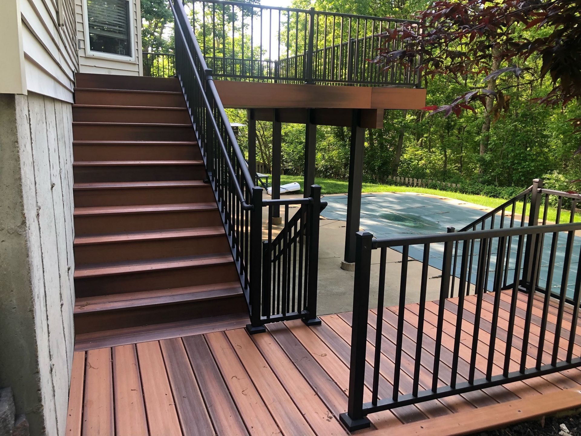 Brown deck and stairs with black railing leading to a raised deck, surrounded by trees.