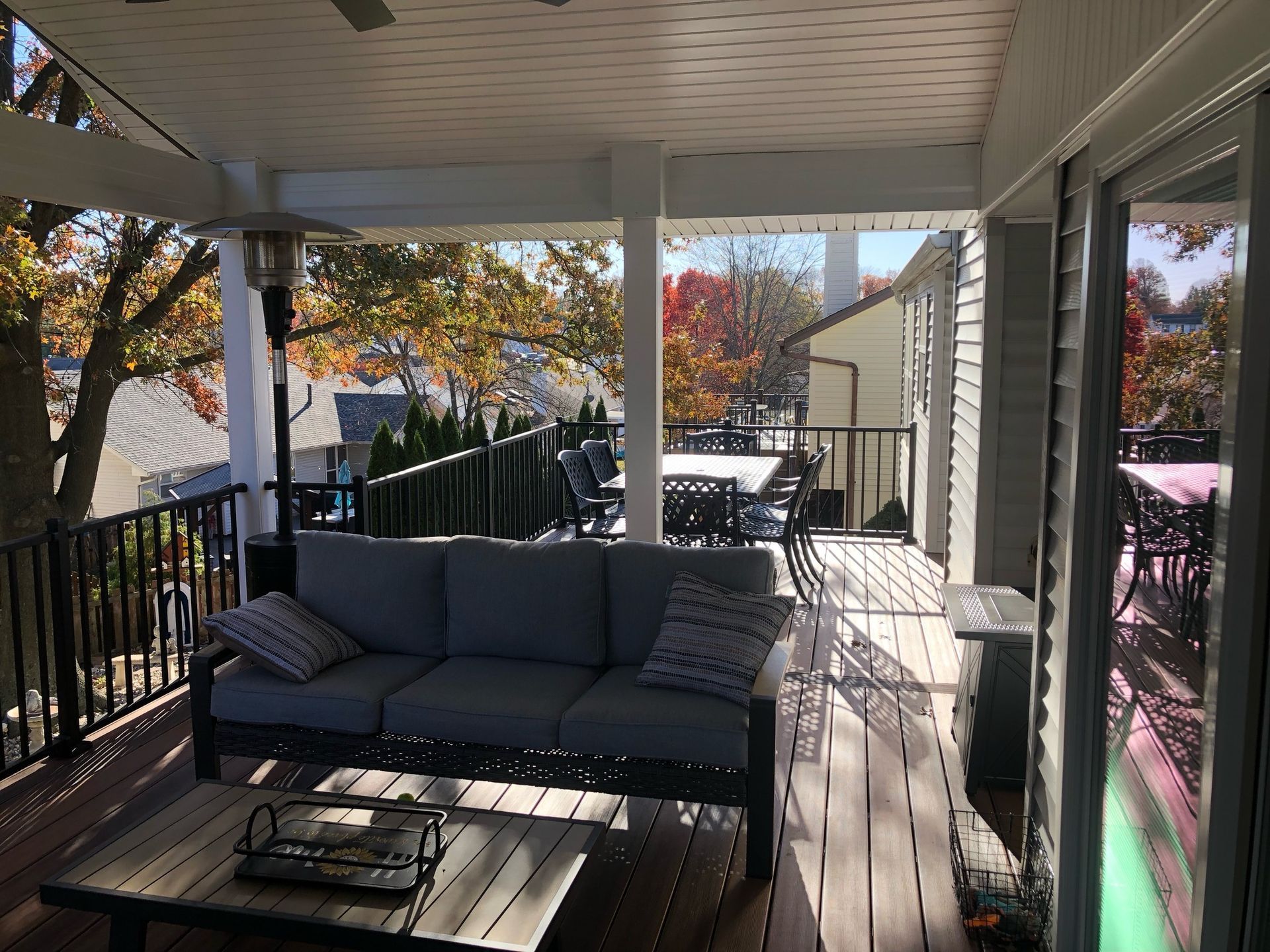 Covered deck with gray sofa, coffee table, outdoor heater, and view of trees.