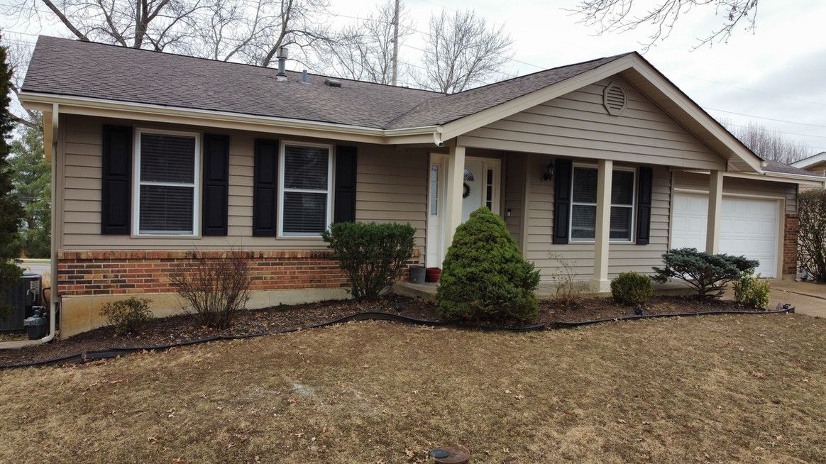 Tan ranch-style house with dark shutters, brick accents, and a two-car garage. Overcast day.