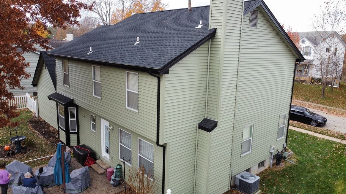 Two-story house with green siding, black roof, and chimney. A patio with furniture is in the backyard.
