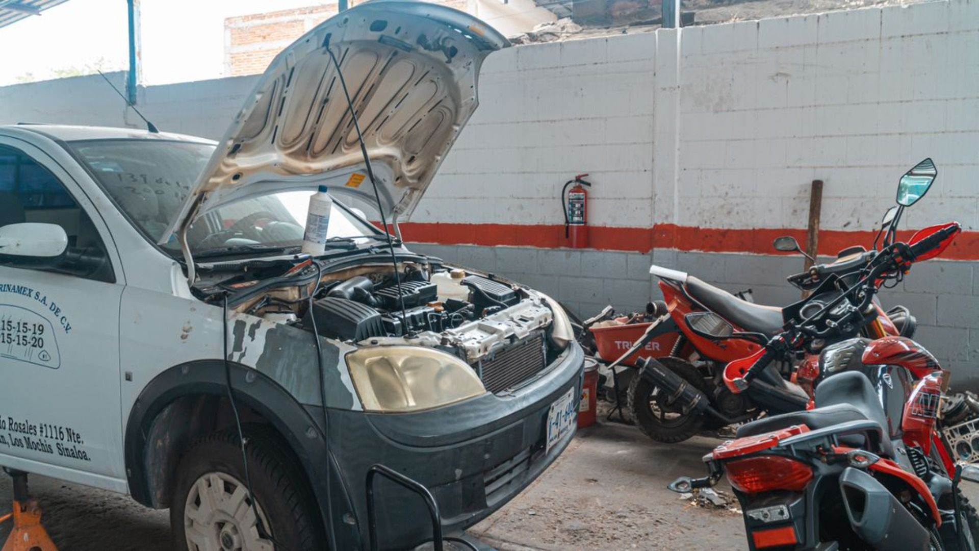 Coche blanco con el capó abierto en un garaje, junto a varias motocicletas aparcadas.