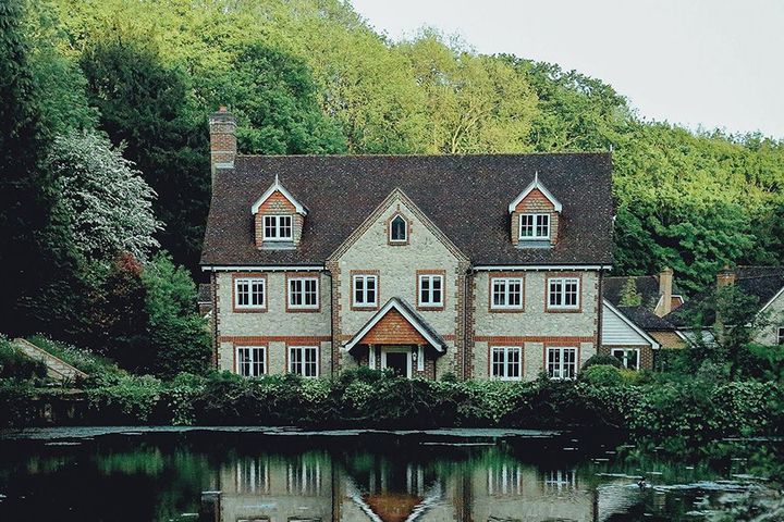 A cozy cottage with red roof and dormer windows reflected in a calm pond, surrounded by trees