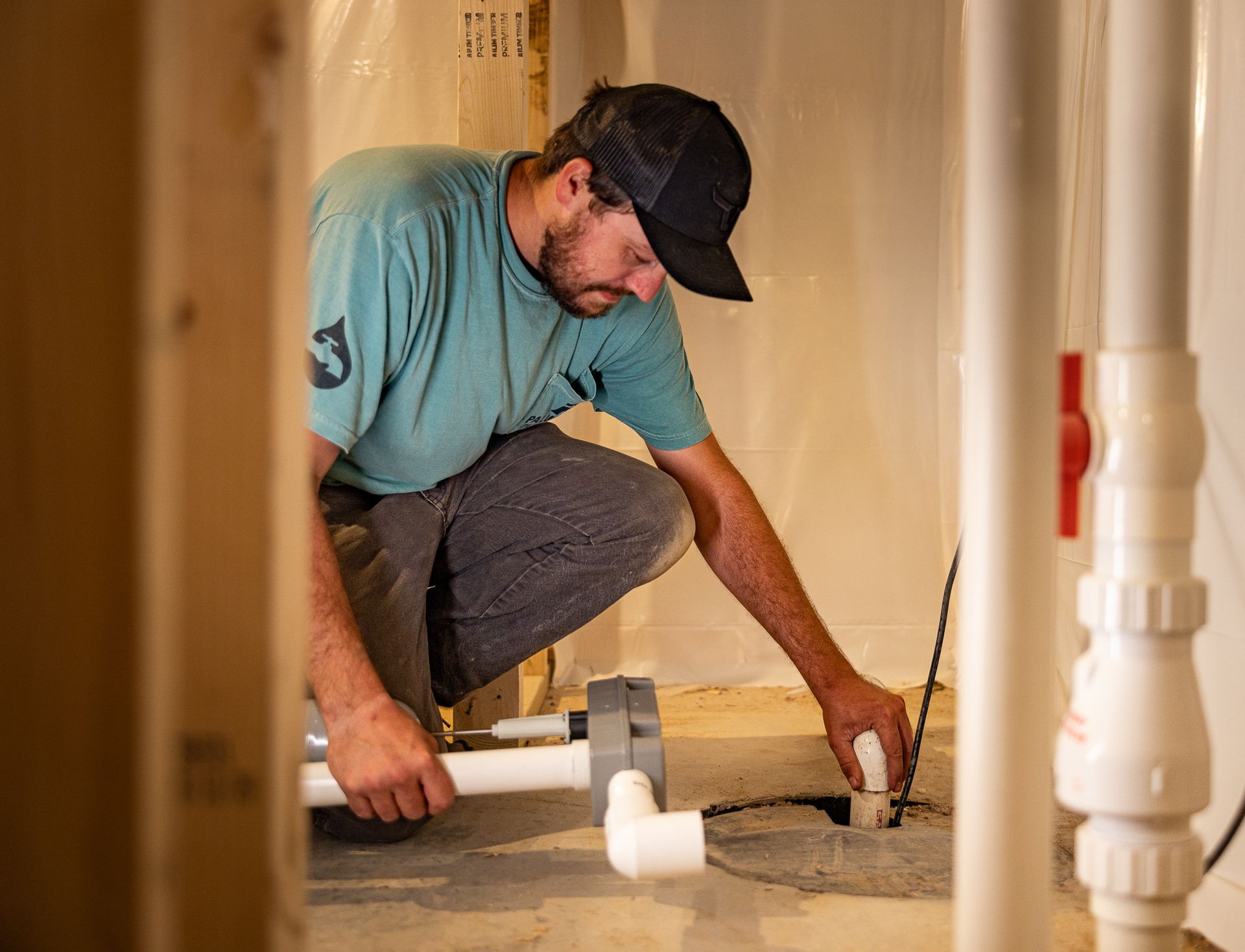 Plumber crouches under a sink, installing white PVC pipes in a narrow hallway.
