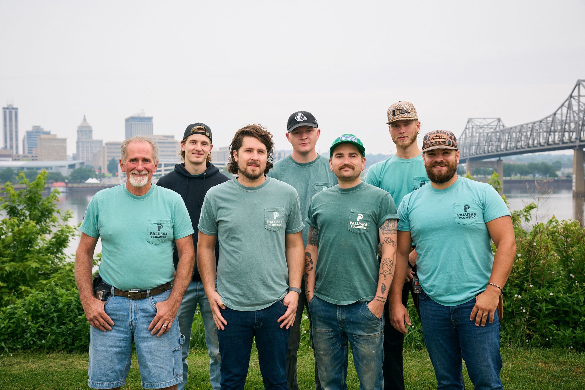 Six men in teal shirts standing outdoors by a river with a city skyline and bridge in the background