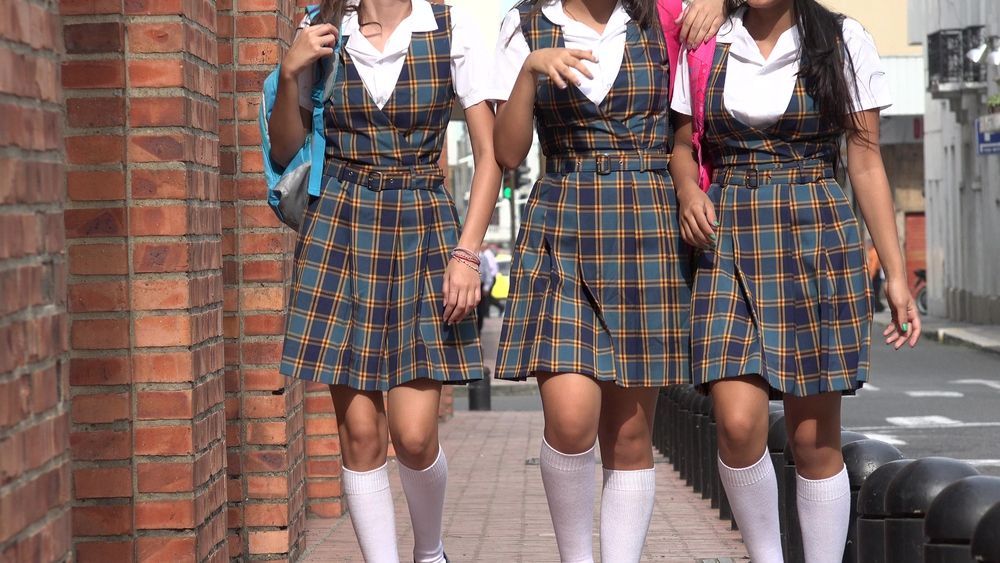 Tres estudiantes con uniformes escolares a cuadros y calcetines blancos hasta la rodilla caminan juntos por una acera junto a un muro de ladrillos.