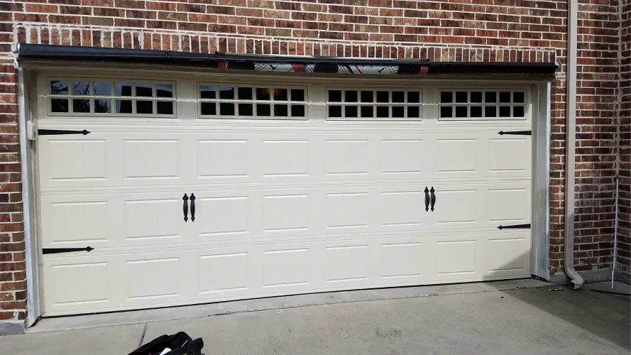 Beige garage door with decorative windows, black hardware, and a brick background.