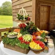 Outdoor buffet table with colorful fruit and vegetable platters beside a wooden building