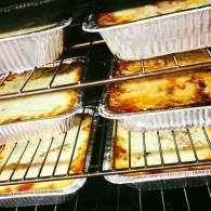 Aluminum trays of baked casserole or lasagna stacked on oven racks under broiling heat