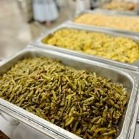 Trays of cooked vegetables in a buffet line, with green beans in the foreground and yellow corn behind.
