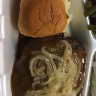 Hamburger patty topped with sautéed onions in a foam takeout container, with the bun above it