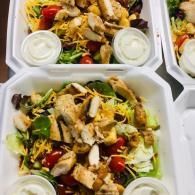 Takeout containers of chicken salad with lettuce, tomatoes, and dressing cups