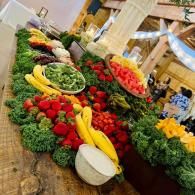 Colorful fruit and vegetable display on a market table, with bananas, strawberries, greens, and melons