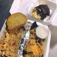 Takeout meal in foam container with pasta, salad, garlic bread, and a dessert cup