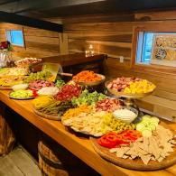 Buffet table with assorted salads, sliced vegetables, meats, and bread in a wood-paneled room