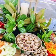 Assorted fresh vegetables and wrapped leaves arranged around a bowl of small stuffed bites on a market stall
