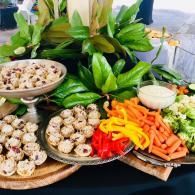 Assorted bite-sized appetizers with vegetables and dip on platters, arranged on a table with green plants in back