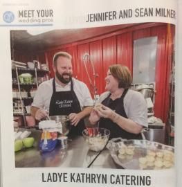 Two people in a kitchen wearing aprons, smiling while preparing food; Lady Kathryn Catering sign visible.