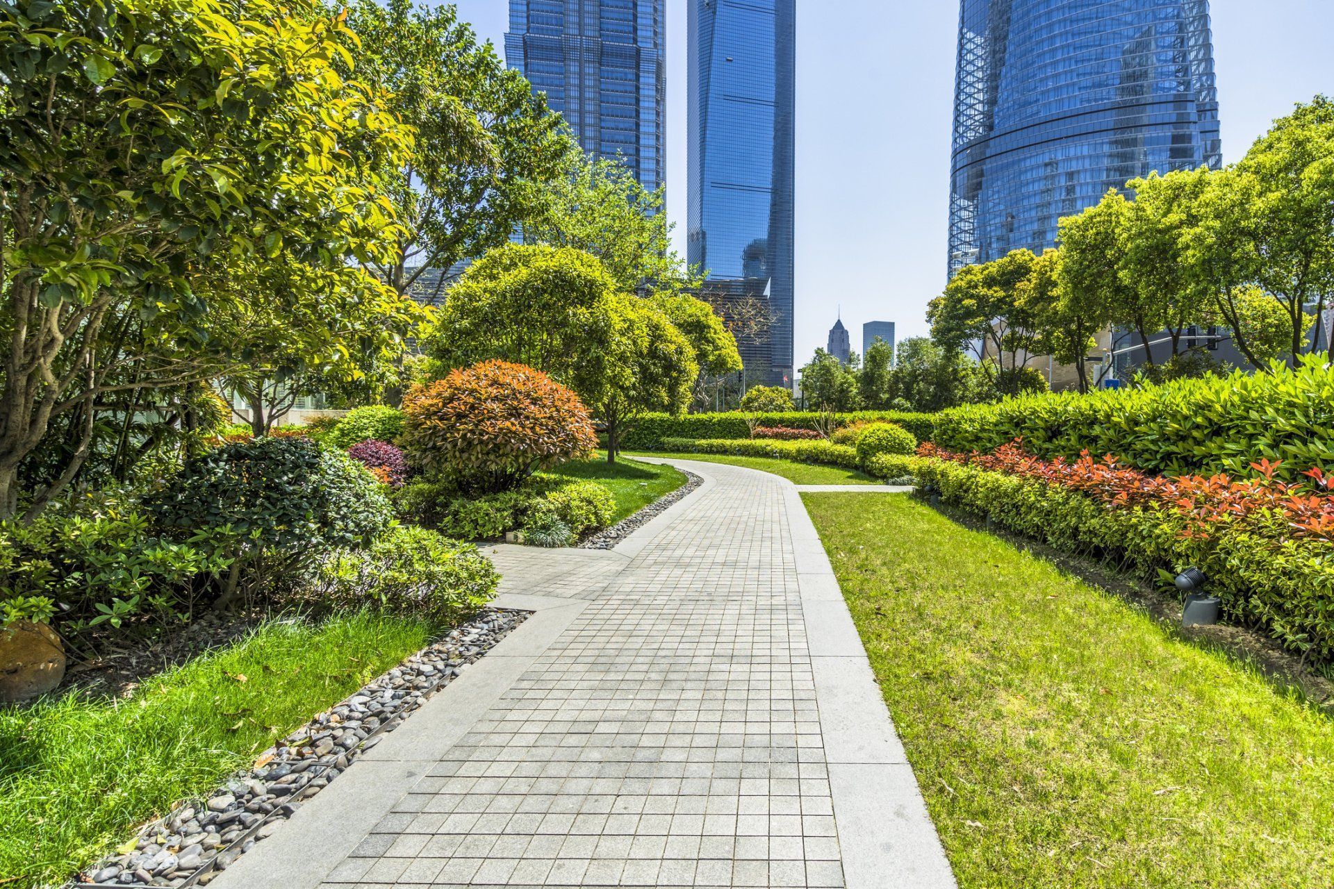 A stone pathway winds through a lush park with green lawns, trees, and shrubs, framed by modern skyscrapers in the distance.