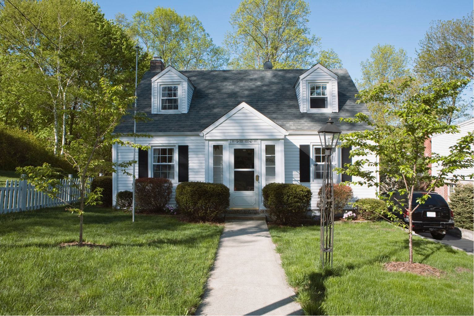 A white house with a gray roof and two dormer windows, surrounded by green trees and a lawn.