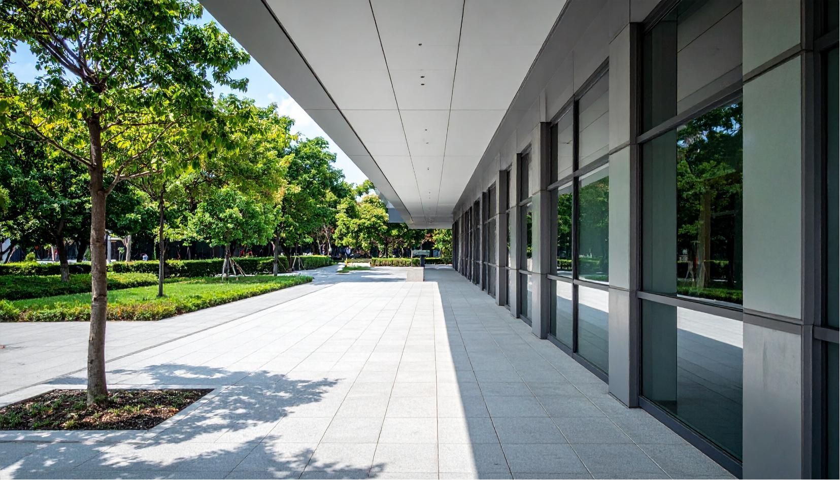A modern building with large glass windows and a wide, covered walkway next to a tree-lined stone courtyard.