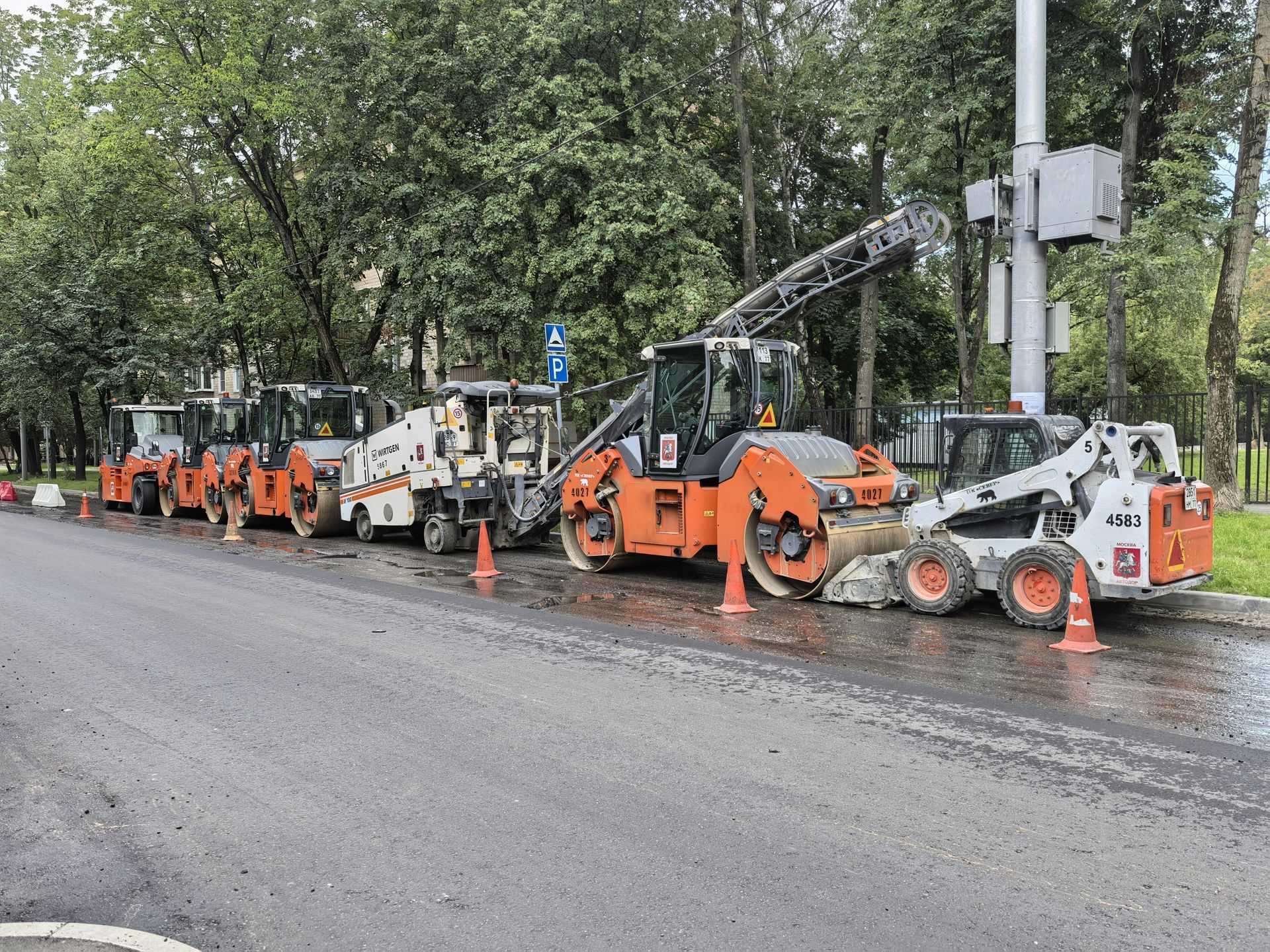 Construction machinery, including rollers and a skid steer, parked along a city road lined with trees.
