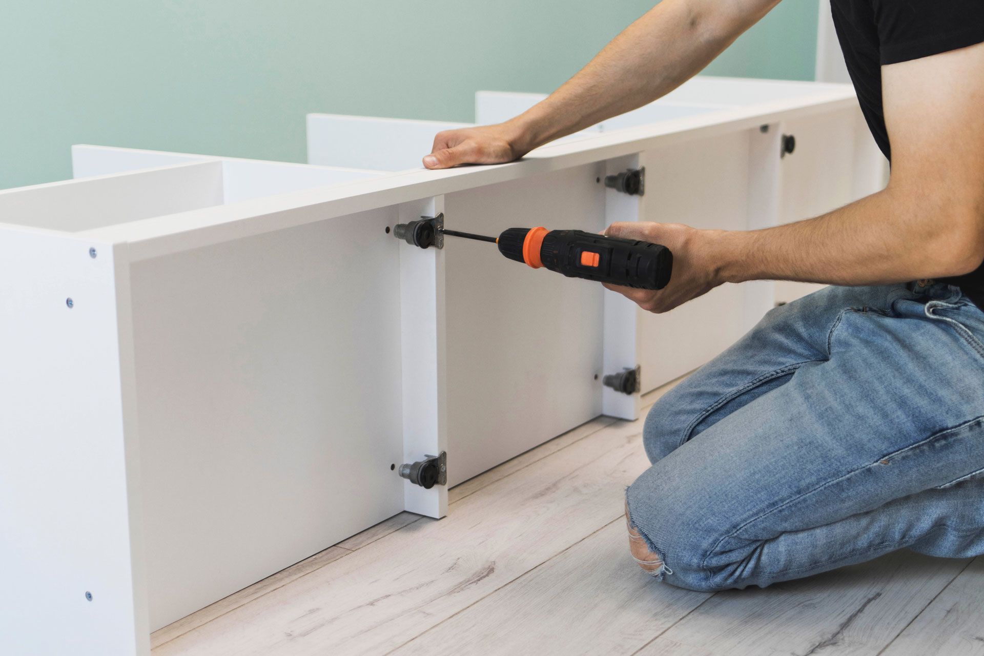 A person kneeling on a floor uses a cordless drill to assemble a white wooden shelving unit.