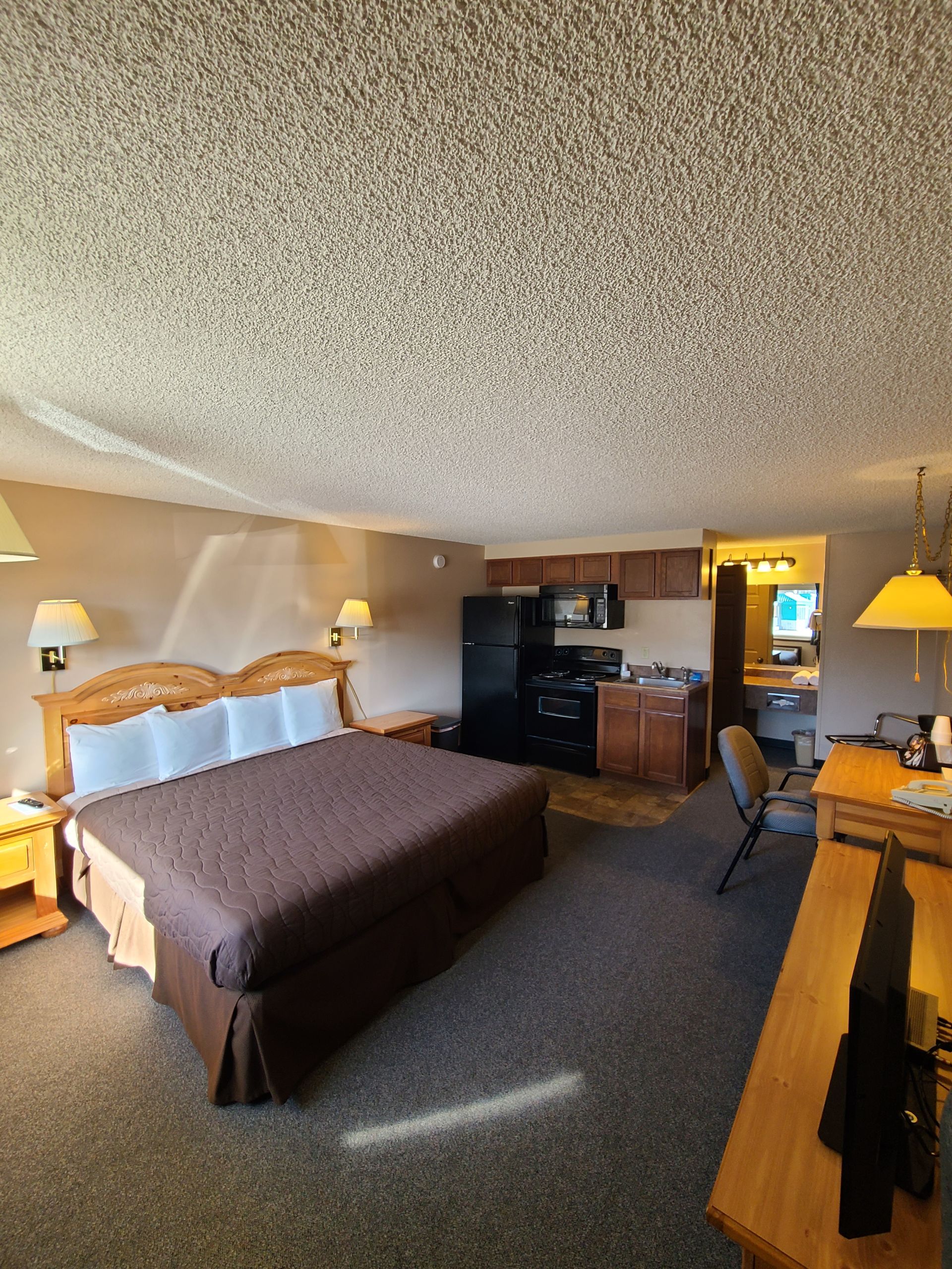 Bedroom of a motel suite with bed, kitchenette, desk, and bathroom.