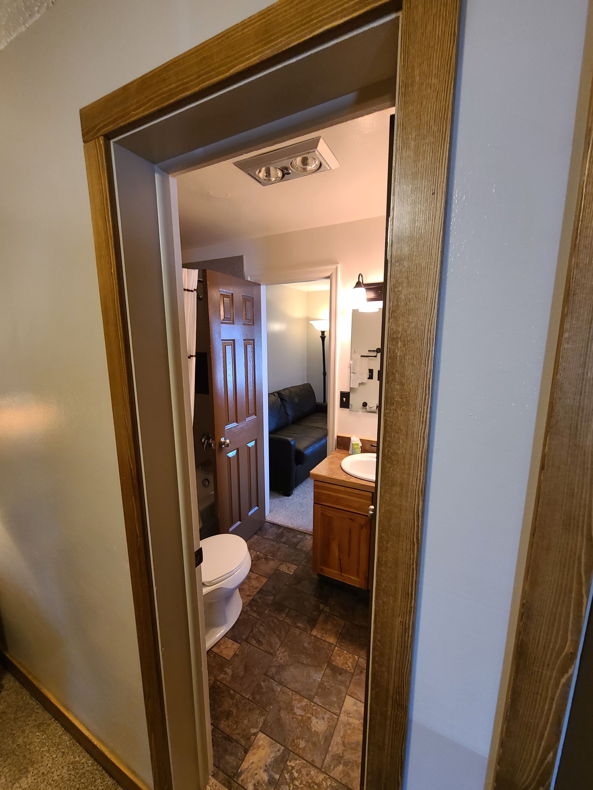 Open doorway into a bathroom with a toilet, sink, and sofa. Brown door frames, wood-look flooring.