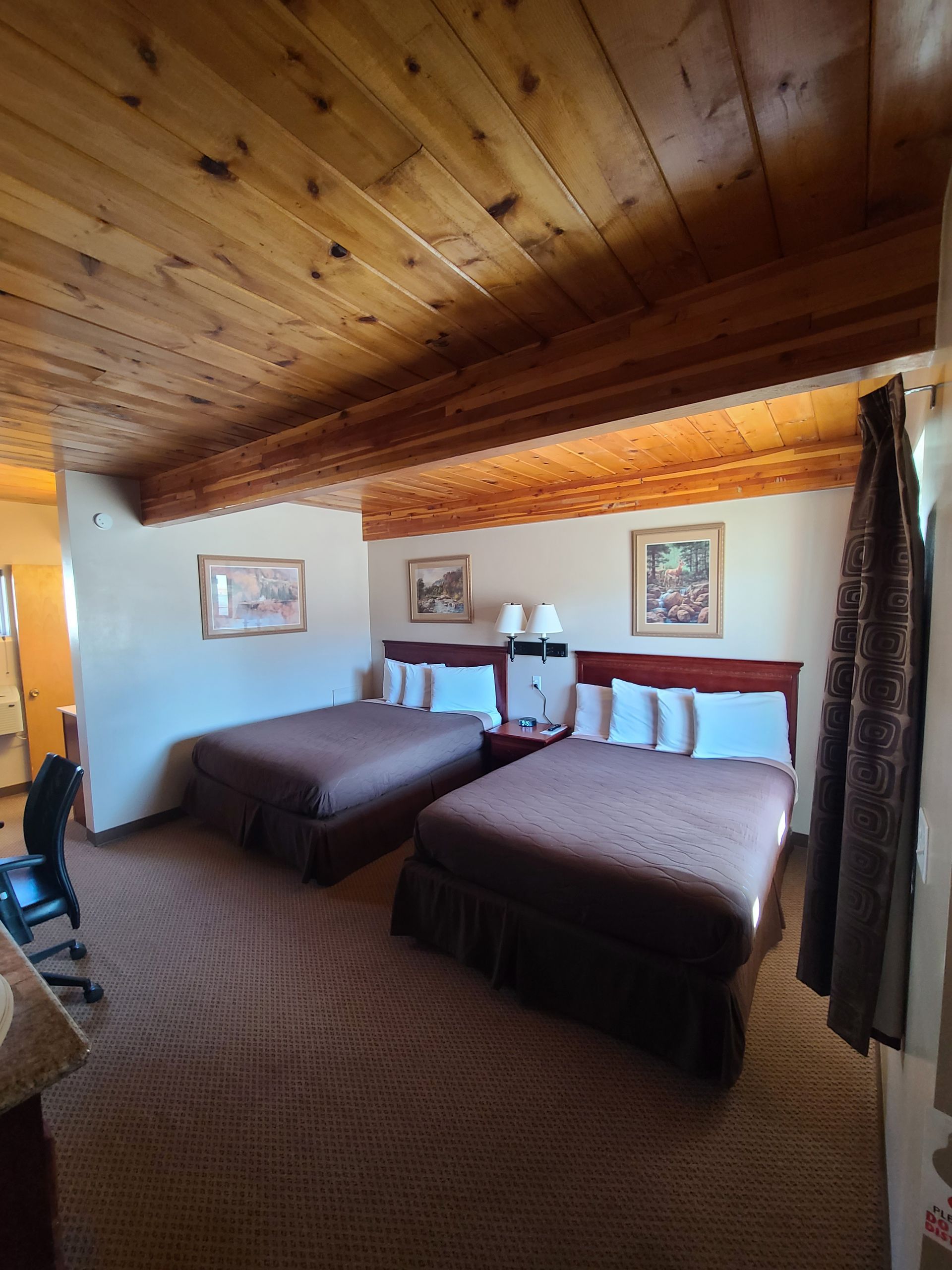 A hotel room with two beds, brown comforters, a wooden ceiling, and a patterned carpet.