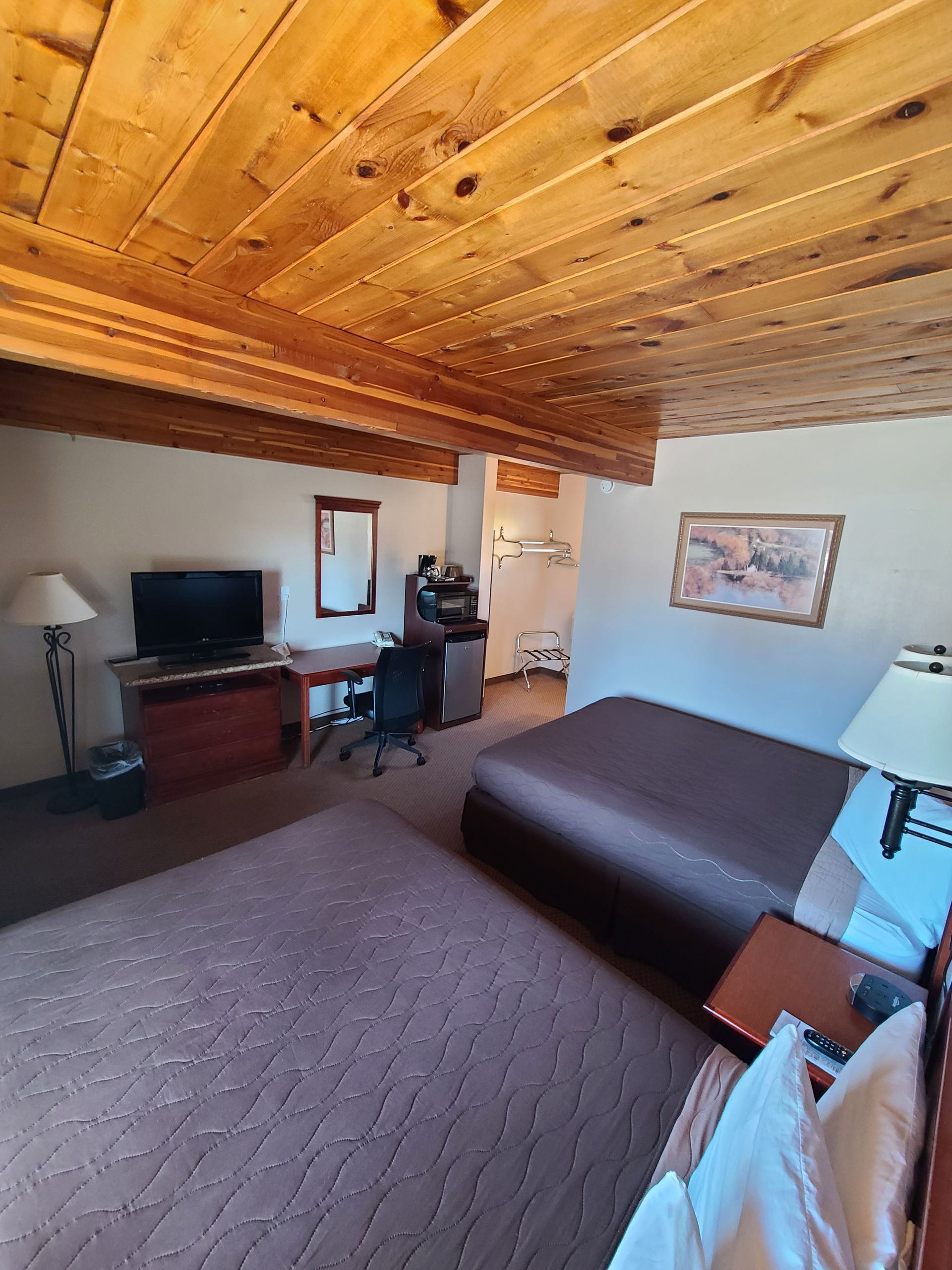 Hotel room with wooden ceiling, TV, desk, bed, and artwork.