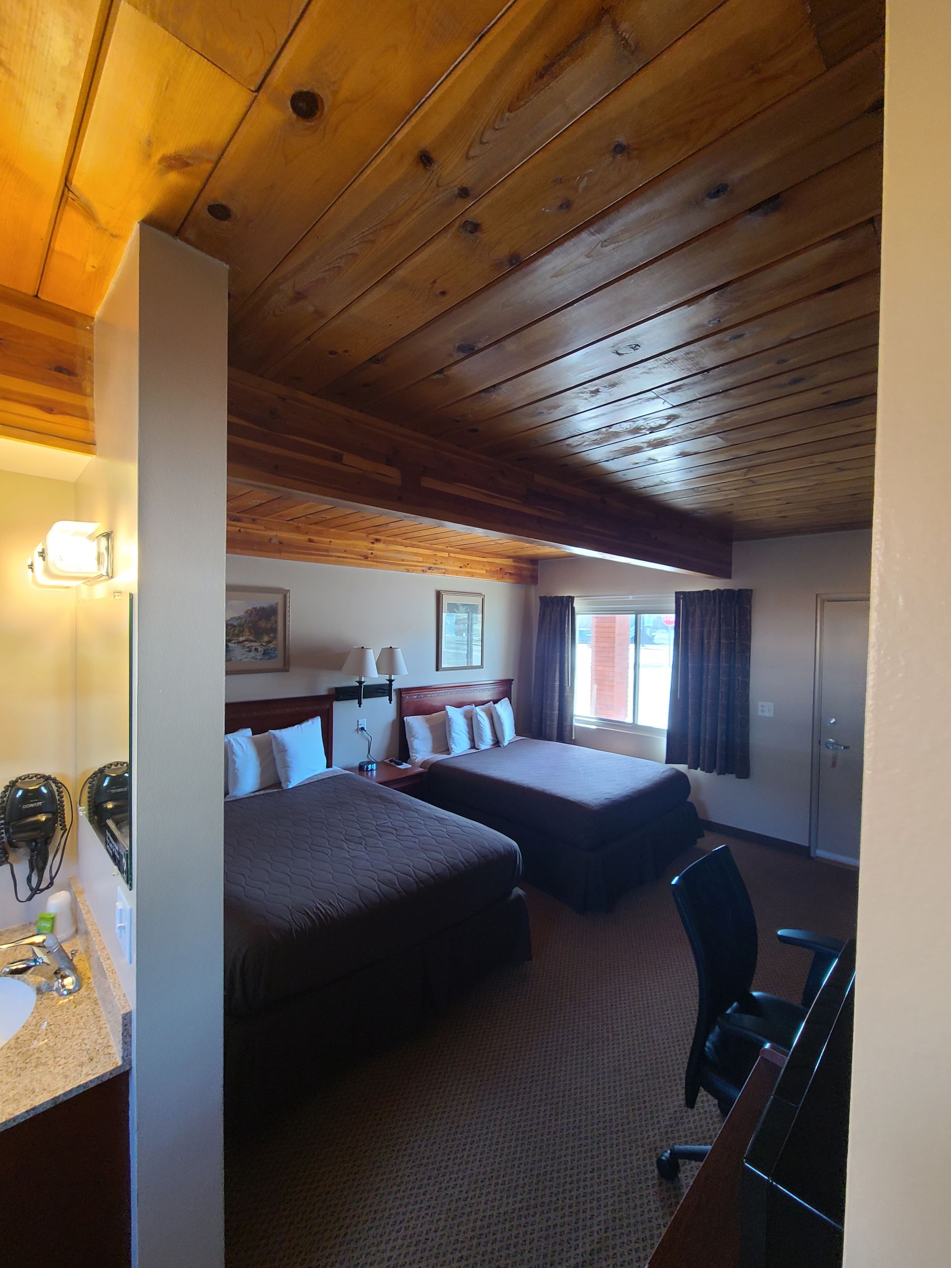 Hotel room with two beds, wooden ceiling, desk, and vanity area. Brown carpet and dark bedding.