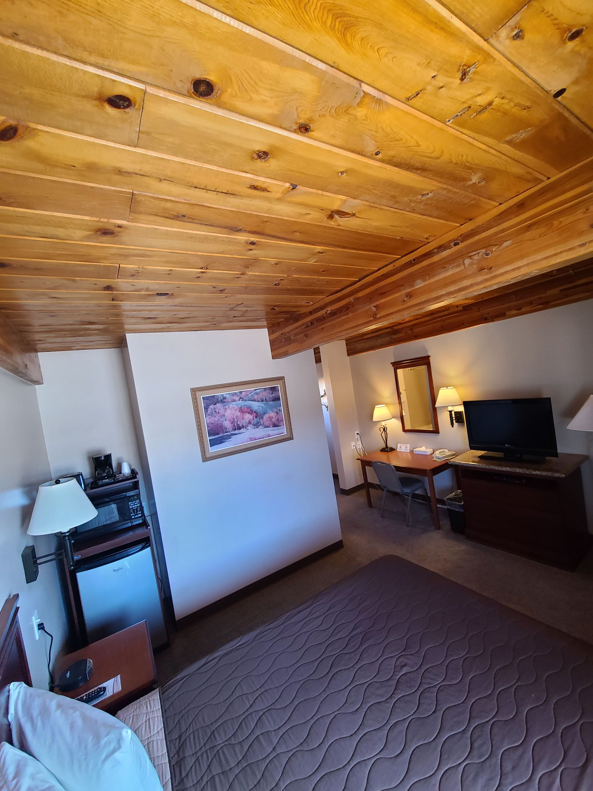A hotel room with a wooden ceiling, bed, TV, desk, and a small refrigerator.