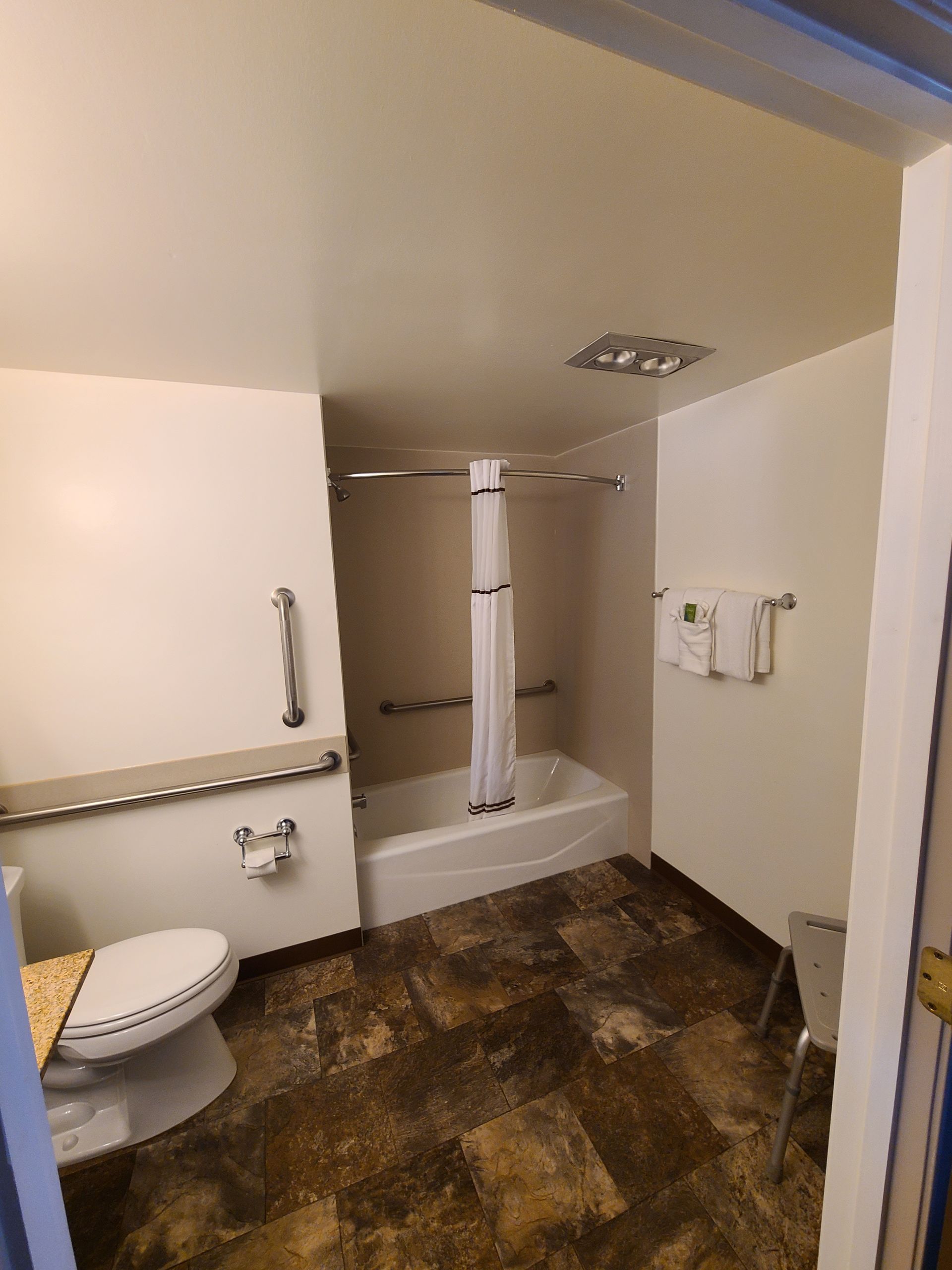 Accessible hotel bathroom: toilet, bathtub with grab bars, towel rack, and brown patterned flooring.