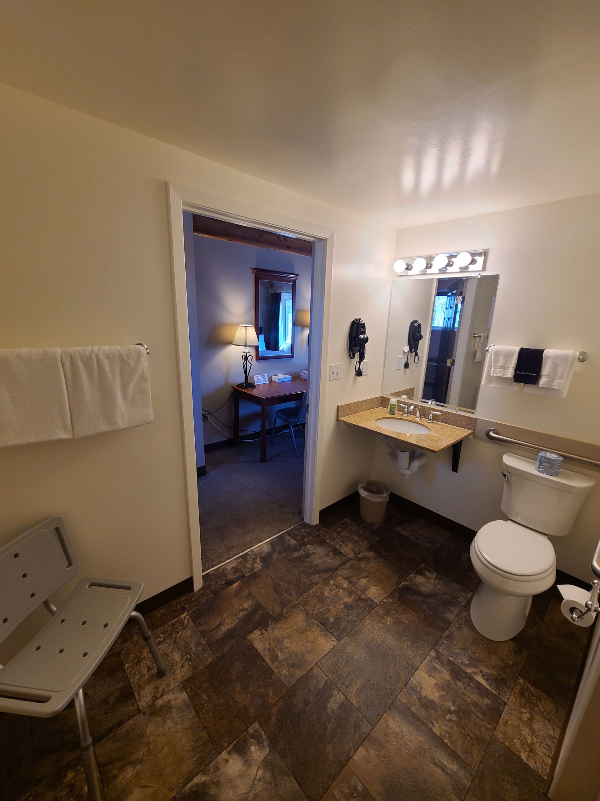 Bathroom with accessible features: open doorway to bedroom, wall-mounted sink, grab bar, shower chair, and toilet.