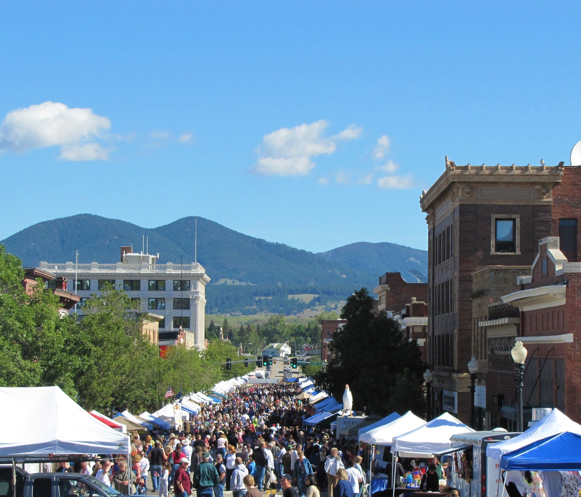 Street fair with crowds, tents, buildings, and mountains in the background under a blue sky.