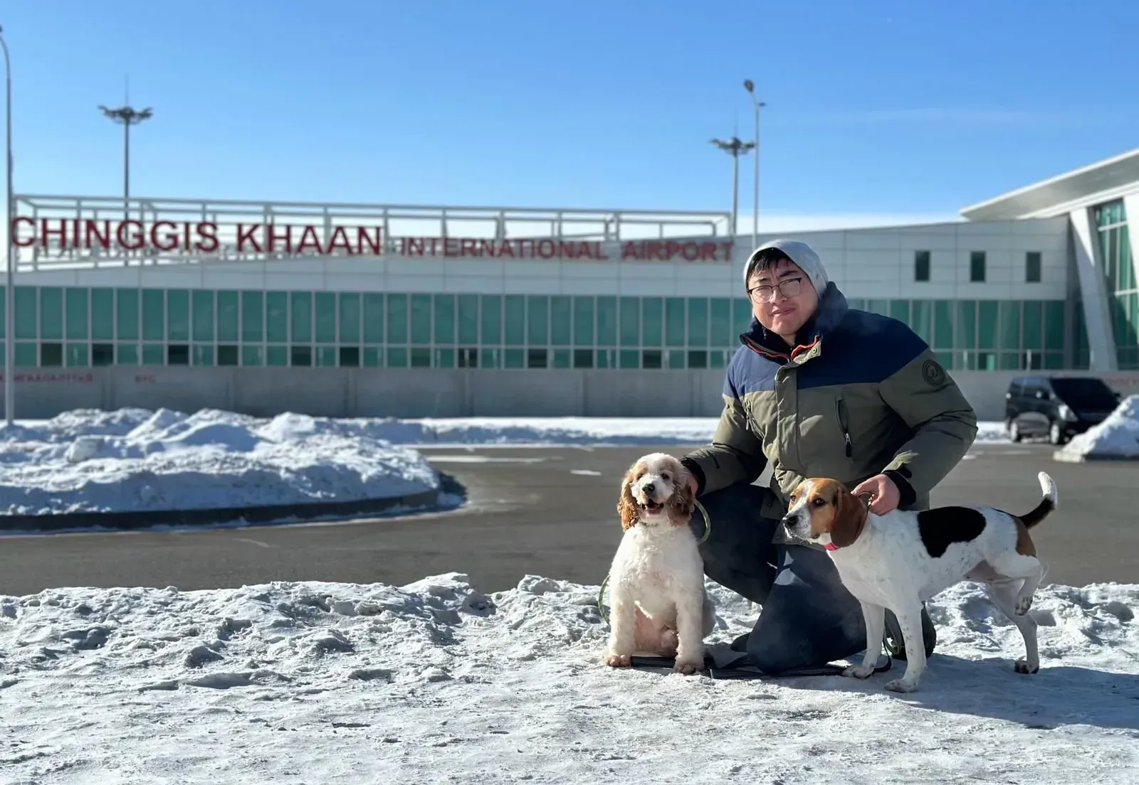 Person crouching with two dogs in front of Chinggis Khaan International Airport on a snowy day