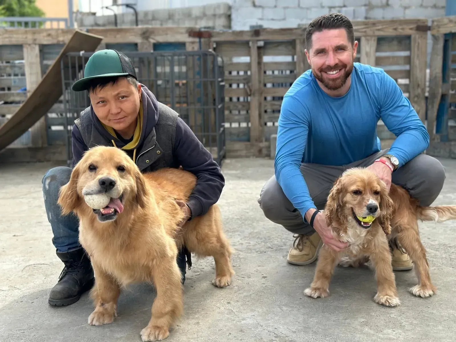 Two men crouch with two golden retrievers in a wooden outdoor area, smiling at the camera.