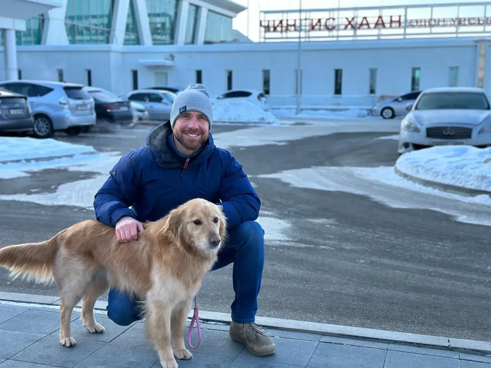 Man crouching beside a golden retriever on a snowy street outside a building