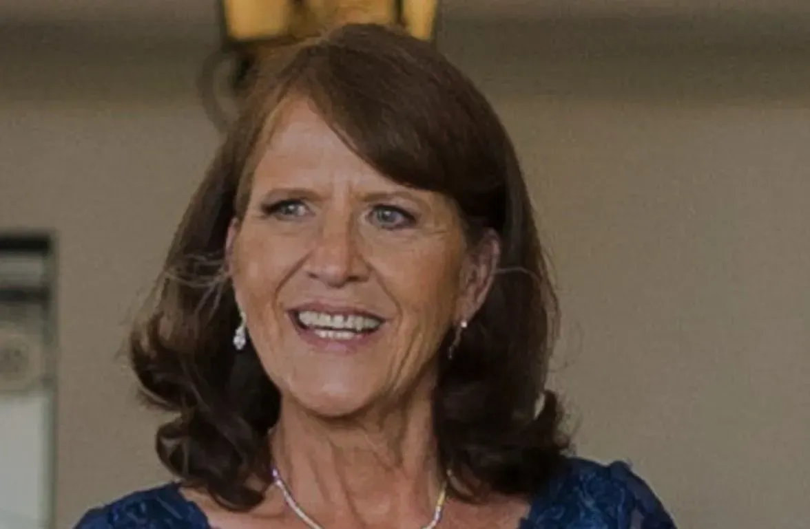 Smiling woman in a dark blue dress and necklace, standing indoors against a blurred background