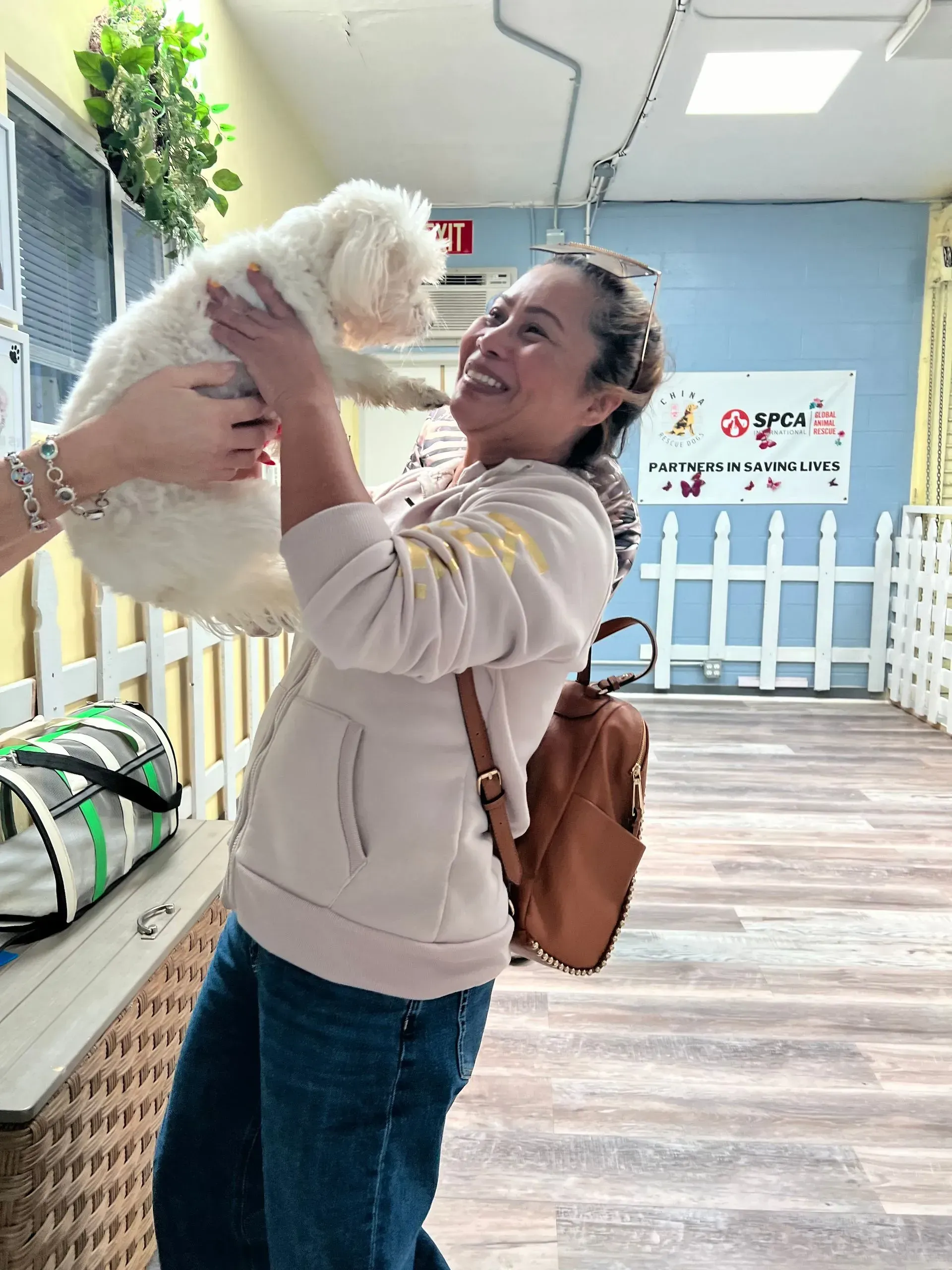Woman holding a small white dog indoors, smiling in a bright pet daycare.