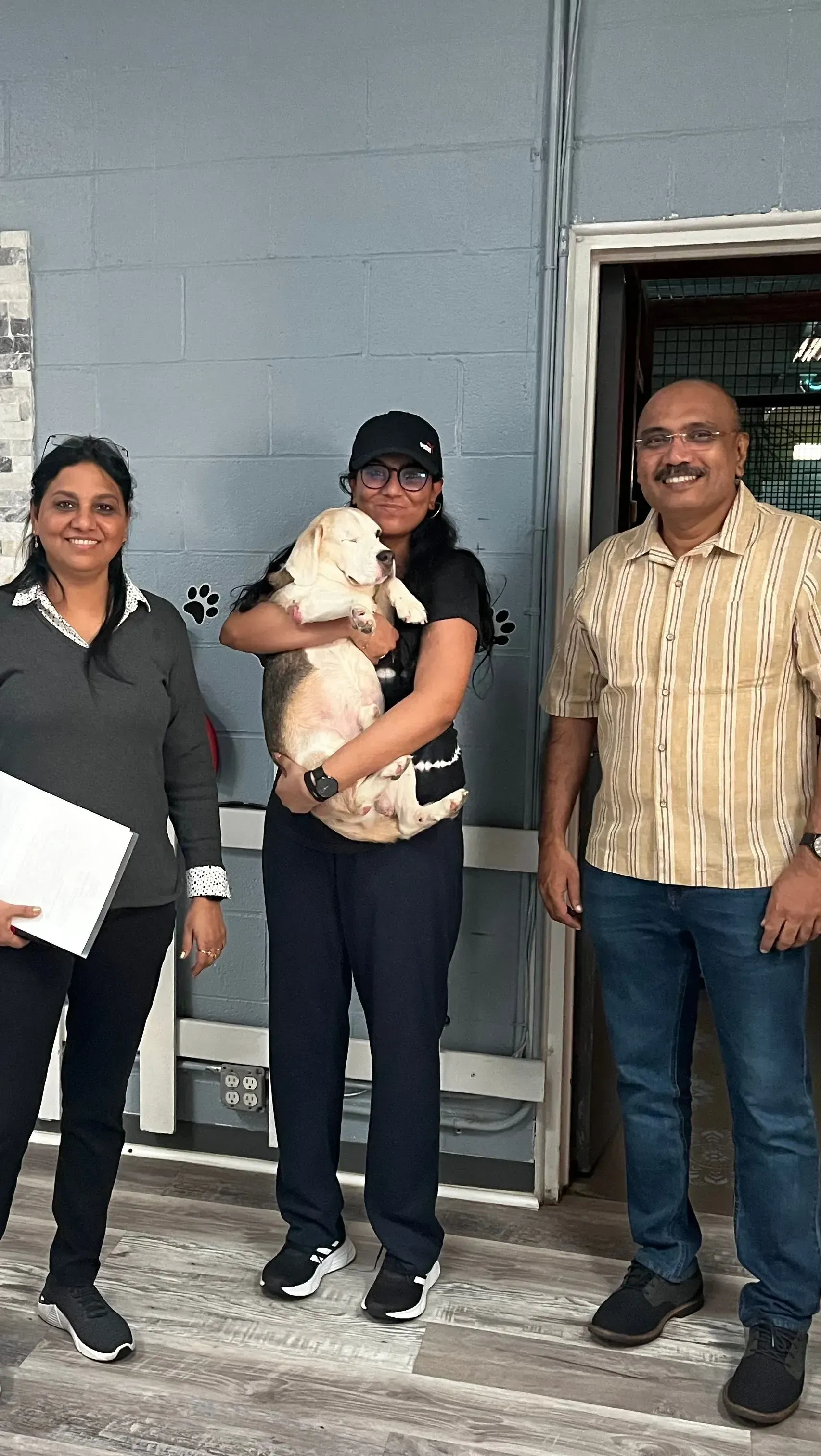 Three people standing by a doorway, one holding a small dog in a gray-walled indoor setting.