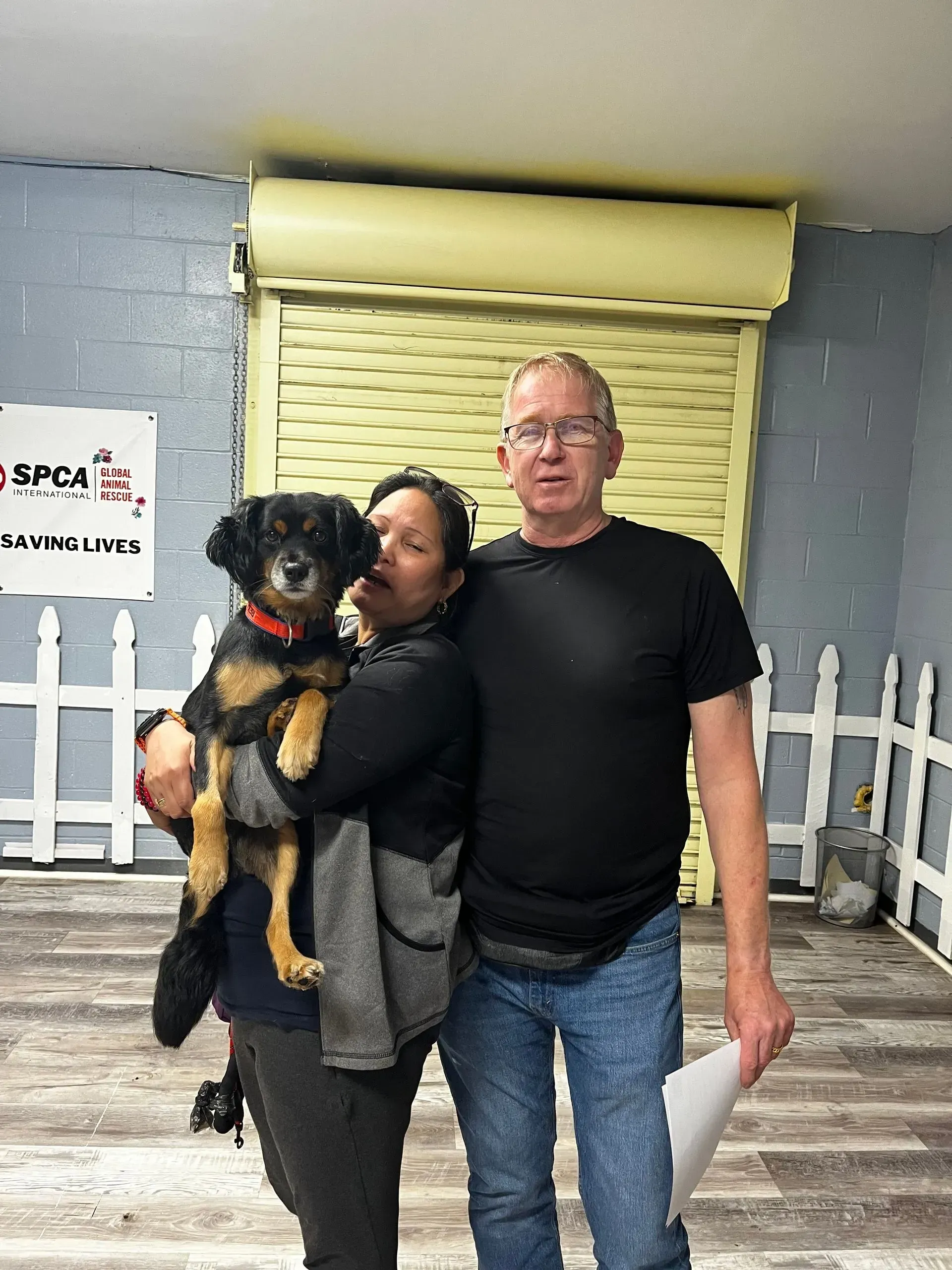 Two people indoors holding a black-and-tan dog, standing by a yellow shutter and white fence backdrop.