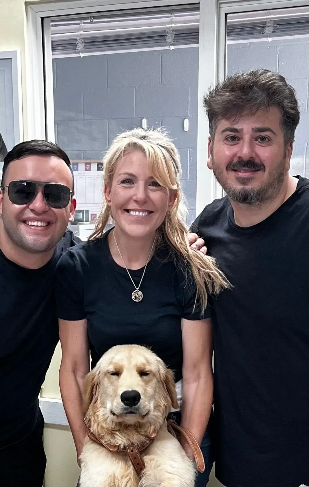 Three smiling people pose indoors with a golden dog in front of a window.