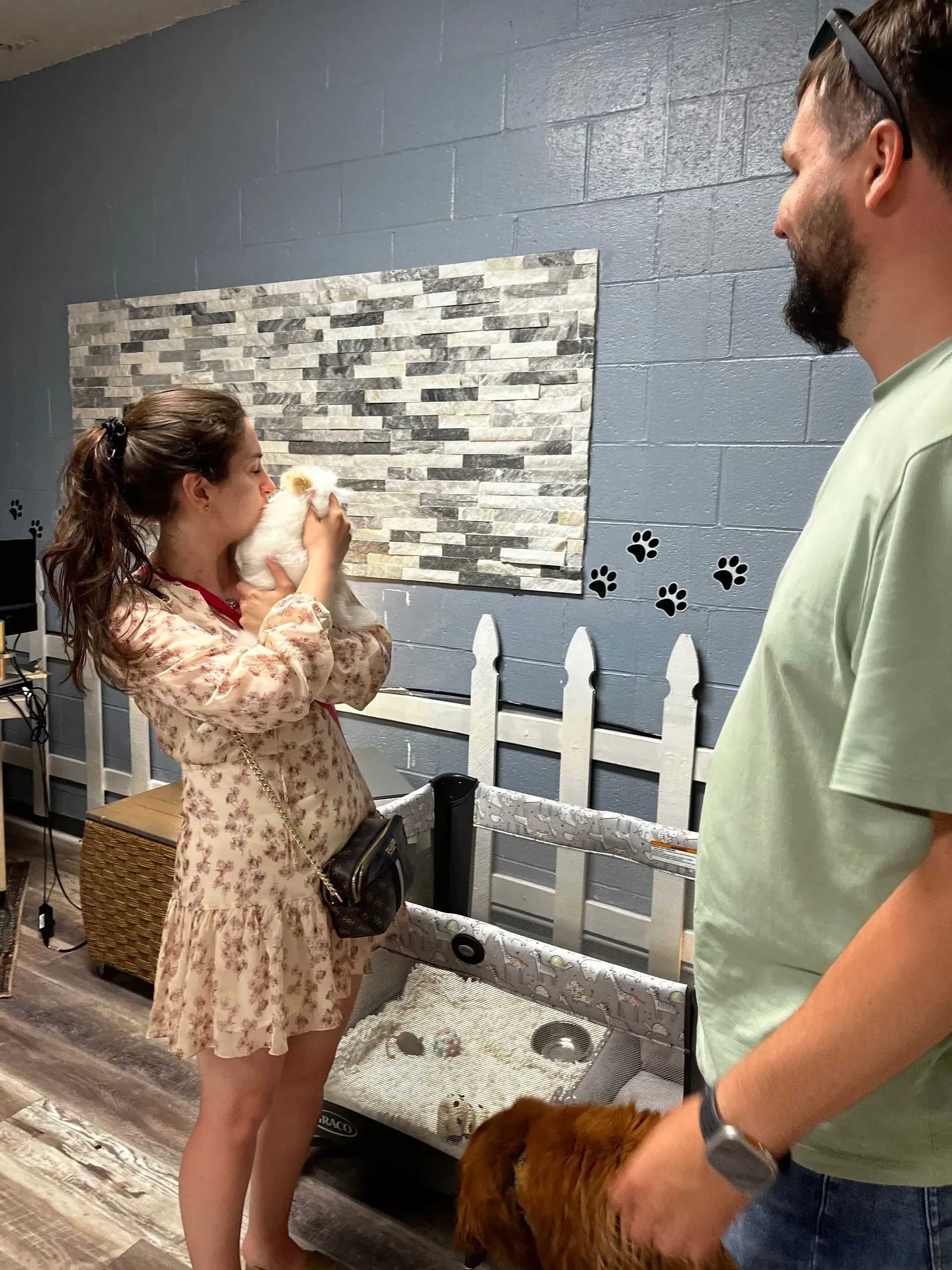 Woman in floral dress taking a drink beside a man in a dog daycare room with white fencing and paw prints