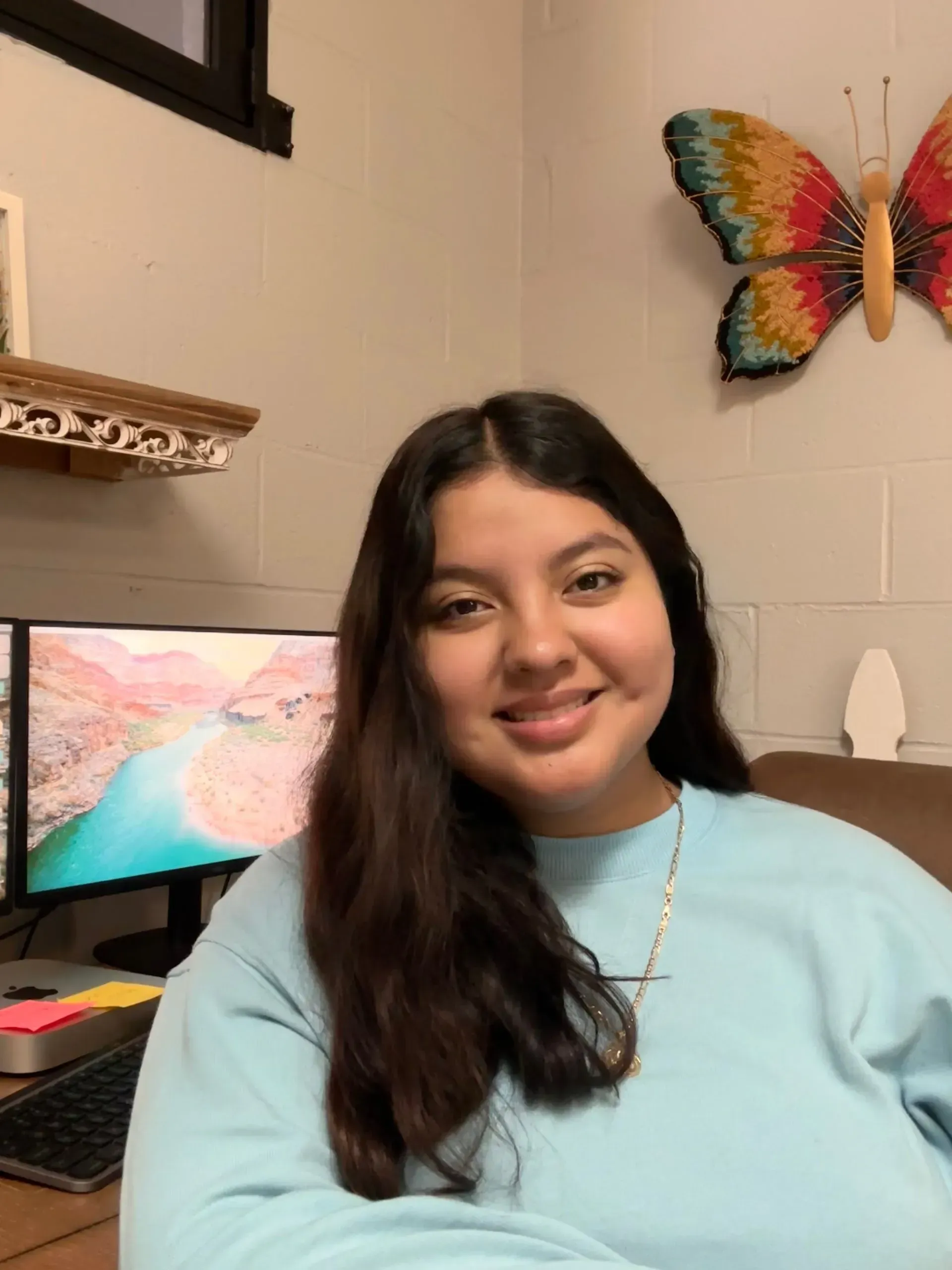 Smiling person in a light blue sweater seated at a desk with a computer and colorful butterfly wall decor.